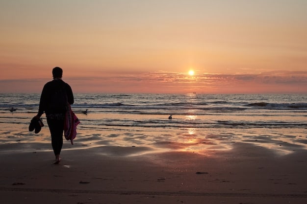 A person walking barefoot on a sandy beach at sunset, with the ocean waves gently lapping at the shore. The sky is a vibrant mix of orange, pink, and purple hues, creating a sense of tranquility and escape.