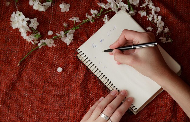 A person's hands carefully sketching in a notebook with a pencil. The notebook is surrounded by art supplies such as colored pencils, erasers, and a small watercolor palette, suggesting a relaxing and creative atmosphere.