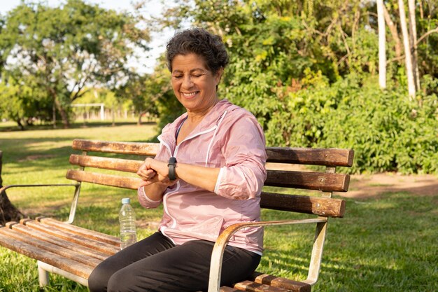 A person using a blood pressure monitor on their arm while sitting on a park bench surrounded by green trees and flowers. They are looking at the monitor with a hopeful expression.