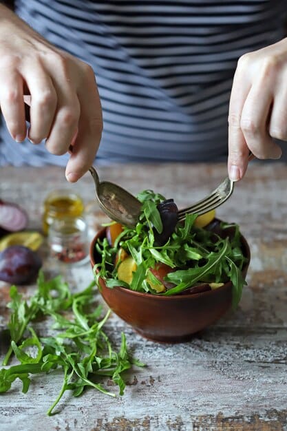 A close-up of a person preparing a colorful salad with fresh, vibrant ingredients, including leafy greens, tomatoes, cucumbers, and bell peppers. The focus is on the act of assembling a healthy, nutritious meal.