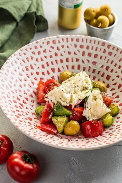 A vibrant, colorful Mediterranean salad in a white bowl, featuring tomatoes, cucumbers, olives, feta cheese, and herbs. Olive oil is drizzled on top. The background is a rustic wooden table with scattered ingredients, creating an inviting and healthy ambiance.