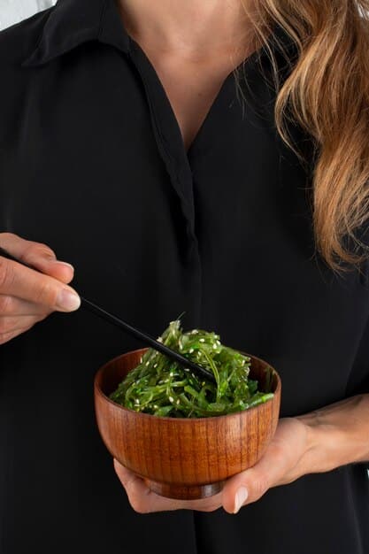 An eye-level, close up shot of a person's hands tossing a vibrant green salad with various vegetables, nuts, and seeds. The person is using wooden salad spoons, and the focus is on the texture and freshness of the ingredients.