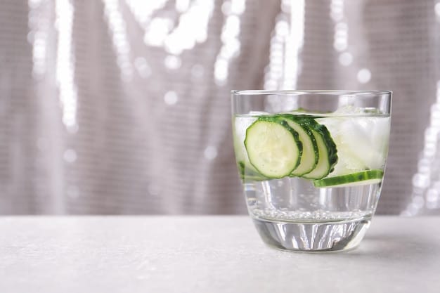 A close-up on a glass of water with sliced cucumber and mint leaves floating inside it. The background should be slightly blurred to put focus on the refreshing drink.