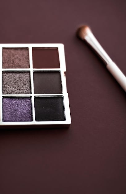 A close-up, top-down shot of a makeup artist's hands applying a dark eyeshadow with a fluffy blending brush onto an eyelid already primed. Various professional brushes and an eyeshadow palette are visible in the background, slightly out of focus.