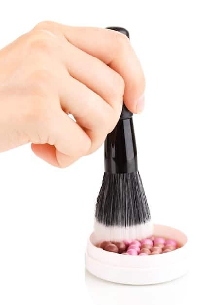 Close-up of a hand holding a makeup brush applying subtle blush to a cheek, showcasing blending technique. The skin looks healthy and radiant.