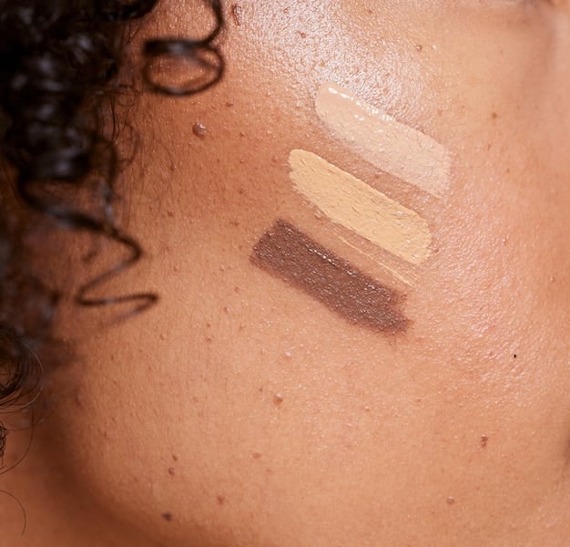 A close-up of a diverse range of liquid foundations and concealers with dewy finishes, some with dropper applicators, alongside blending sponges and brushes on a light background, emphasizing natural, light-reflecting makeup.