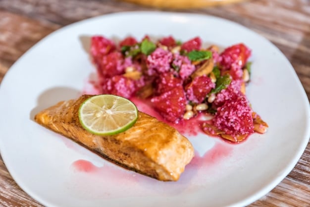 A close-up shot of a healthy grilled salmon fillet, flaked to show its rich, pink, moist texture, alongside fresh sprigs of dill and lemon wedges on a rustic wooden board. The scene emanates freshness and nutritious appeal.