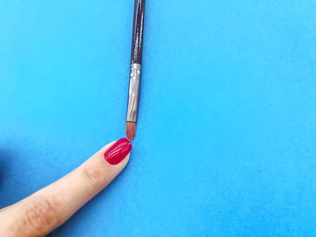A manicurist's hand holding a fine-tip nail art brush, carefully applying a thin black line over a pastel blue nail, with various nail art tools scattered in the background.