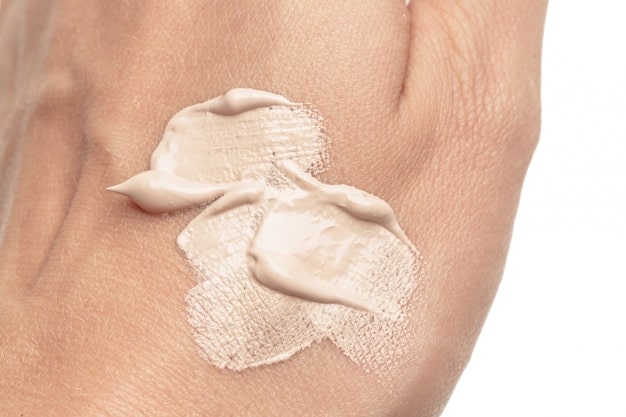 A close-up of a hand applying a pea-sized amount of tinted moisturizer to the face, showing a natural, glowing skin effect.