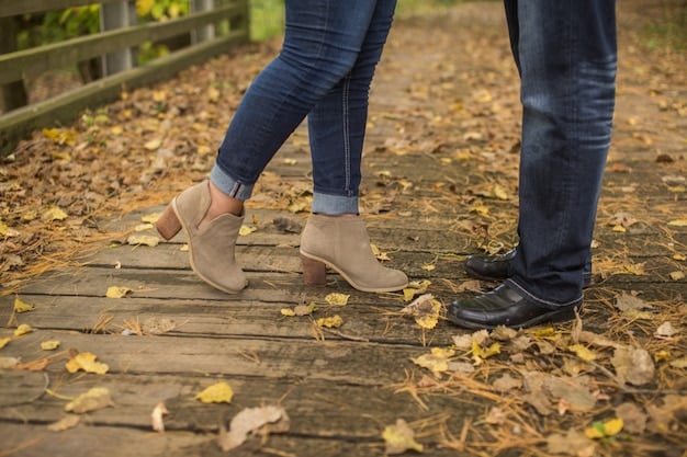 A close-up of a person's feet on a sidewalk in autumn, wearing stylish, dark olive-green suede ankle boots with a low block heel, paired with cuffed dark wash jeans. Leaves are scattered around the boots.