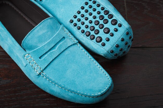 A pair of elegant, low-heeled mules made of soft, perforated suede in a pastel blue, featuring a unique, sculptural transparent heel, resting on a clean, light-colored wooden floor bathed in soft morning light.