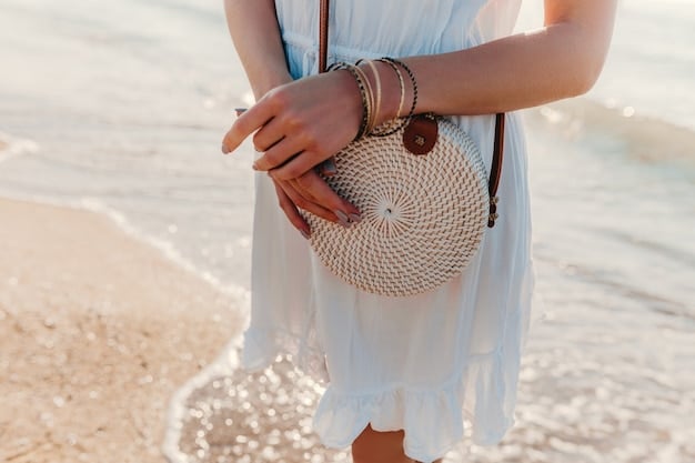 A close-up of a stylish woman's hand adorned with delicate gold bracelets and rings, holding a woven straw clutch bag, with a sun-drenched background indicating summer.