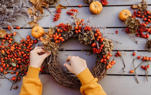 Hands crafting a fall wreath with colorful faux leaves, berries, and a small decorative pumpkin attached to a grapevine base. The background shows other craft supplies.