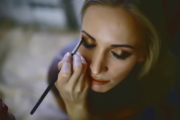 A makeup artist applying dark eyeshadow to the outer corner of a woman's eye with a blending brush. The focus is on the blending technique, demonstrating how to create a gradient effect. Other essentials like makeup palettes and additional brushes are in the background. This illustrates Step 3 of the Smokey Eye Tutorial: A Step-by-Step Guide to a Classic Evening Look - Adding Depth with a Darker Shade.