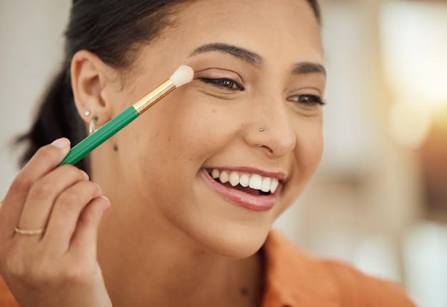 A woman applying liquid concealer under her eyes with a small brush, focusing on brightening the area. The background is a professionally lit studio setting, highlighting the smooth application and skin tone match as part of 'The Ultimate Guide to Concealer: Brightening, Correcting, and Covering'.