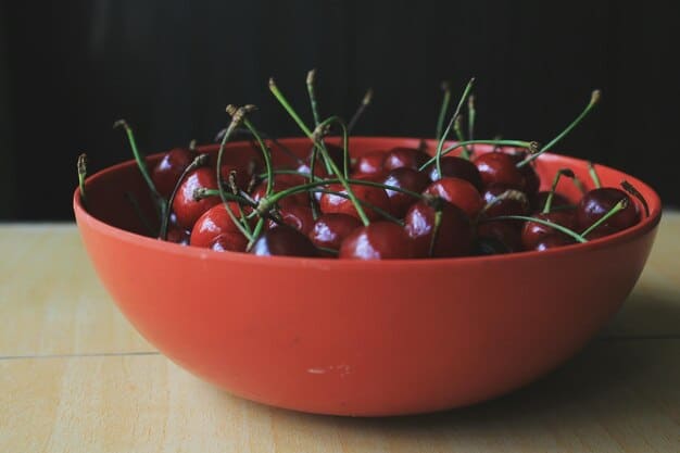 A close-up of a bowl filled with ripe cherries. The cherries are deep red and glistening, with a few still attached to their stems. The background is softly blurred, highlighting the freshness and appeal of the fruit.