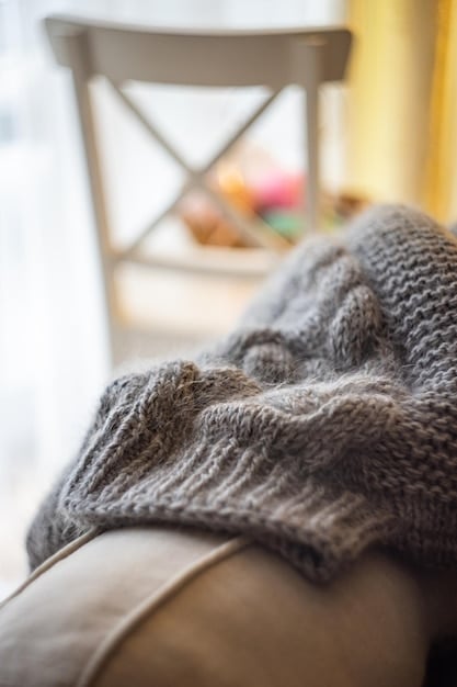 A close-up shot of a textured throw blanket draped over a chair. The blanket is made of a chunky knit material in a neutral color, like cream or beige. The texture is highly visible and tactile, creating a sense of warmth and comfort. The chair is partially visible, suggesting a cozy reading nook or seating area. The focus is on the texture of the blanket and its contribution to a cozy fall atmosphere, referencing Fall Home Decor: Transform Your Living Space with 8 Cozy and Budget-Friendly Seasonal Updates.