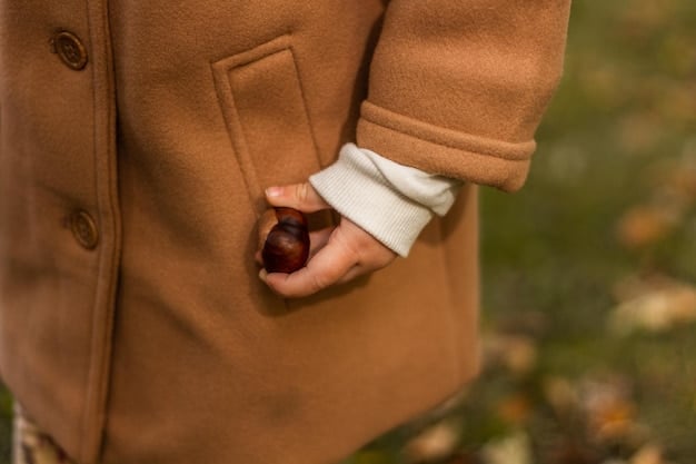 A close-up shot of a woman's hand adjusting the collar of a brown wool coat. The coat has a subtle herringbone pattern and is accessorized with a colorful scarf. The background is blurred, emphasizing the texture and detail of the coat.