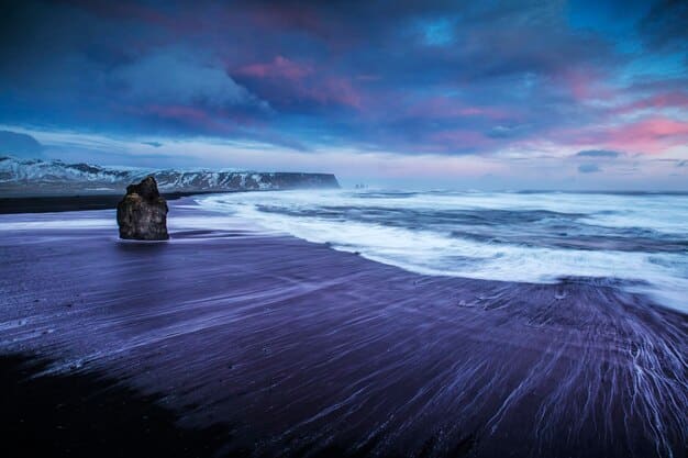 A serene beach scene in Iceland, with black sand, clear blue water, and distant mountains partially covered in snow. The image highlights the unique and cool environment of Iceland, ideal for Summer Travel: Top 3 Destinations for a Relaxing Getaway and Beat the Heat in 2025.